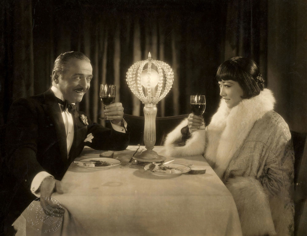 A promotional still for Pavement Butterfly showing Gaston Jacquet and Anna May Wong, seated at a dining table, raising their glasses to toast.